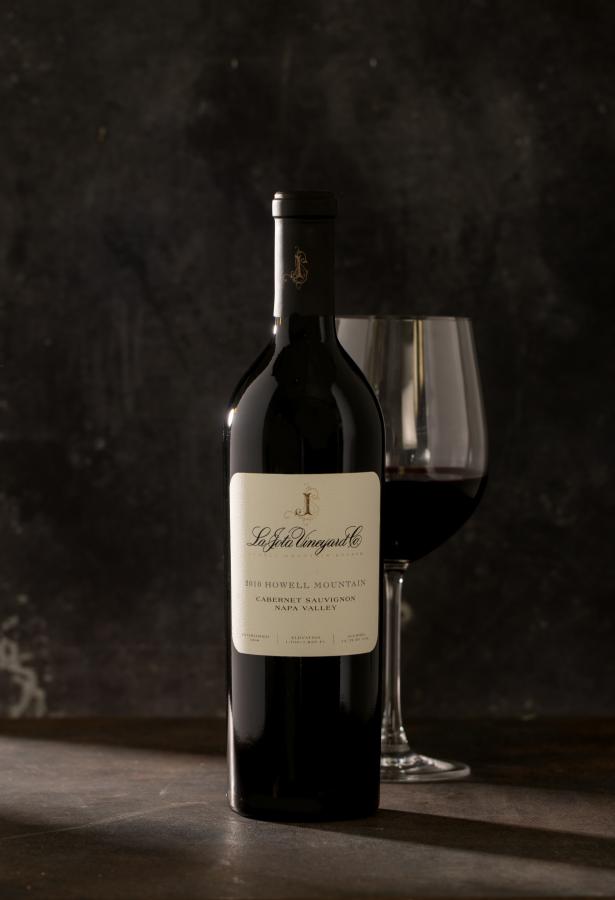 Single bottle of red wine with a wine glass behind the bottle against a dark gray background.