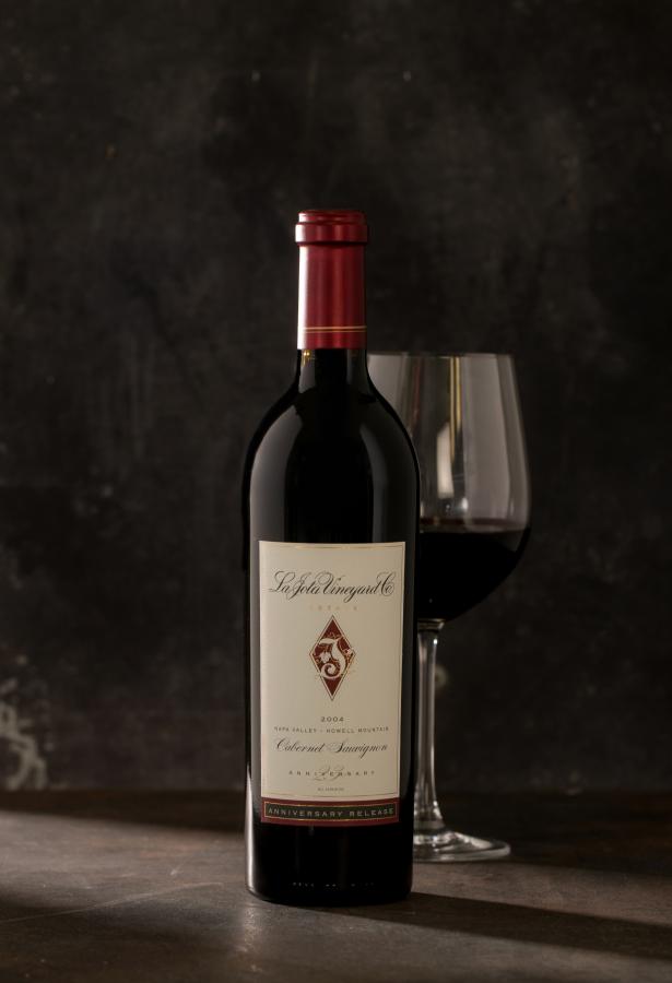 Single bottle of Cabernet with a wine glass behind the bottle against a dark gray background.