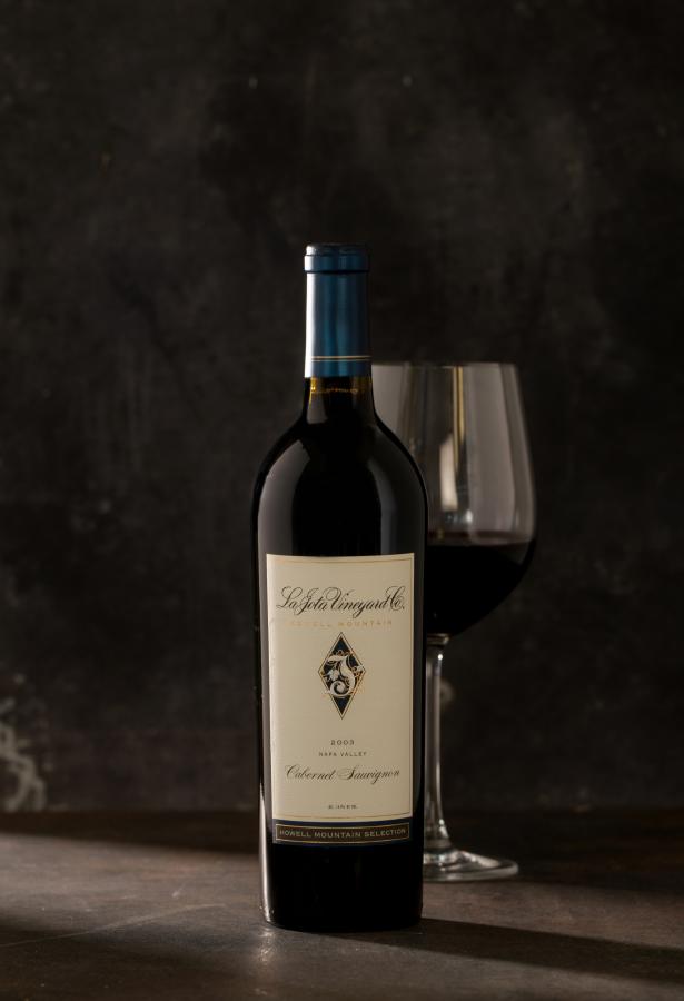 Single bottle of red wine with a wine glass behind the bottle against a dark gray background.