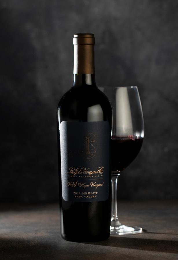 Single bottle of red wine with a wine glass behind the bottle against a dark gray background.